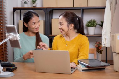 Başarı ve neşeli, girişimci veya çevrimiçi satıcı, ecommerce veya startup business, küçük işletme ile mağaza incelemesi için tablet ve dizüstü bilgisayar kullanan iki genç Asyalı kadın.