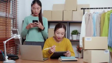 Tablet üzerinde çalışan iki genç Asyalı kadın çevrimiçi iş yaparken ve planlarken ve akıllı telefondaki konuşmayla siparişleri kontrol ederken, çevrimiçi satıcı, girişim şirketi ve kargoyla perakende satış yaparken.