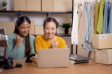 Başarı ve neşeli, girişimci veya çevrimiçi satıcı, ecommerce veya startup business, küçük işletme ile mağaza incelemesi için tablet ve dizüstü bilgisayar kullanan iki genç Asyalı kadın.