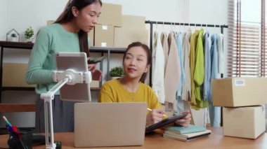 Tablet üzerinde çalışan iki genç Asyalı kadın çevrimiçi iş yaparken ve planlarken ve akıllı telefondaki konuşmayla siparişleri kontrol ederken, çevrimiçi satıcı, girişim şirketi ve kargoyla perakende satış yaparken.