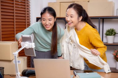 İki genç Asyalı kadın online giyim mağazasında yaşarken, evde sosyal medyada tablet ve dizüstü bilgisayarlı giysiler gösteriliyor. Girişimci çevrimiçi kıyafet satışı ve sunumu için çevrimiçi satış yapıyor..