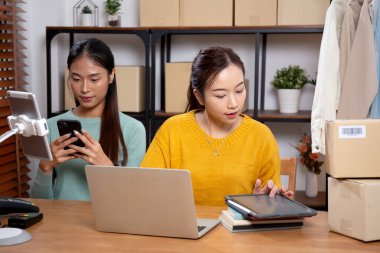 Tablet üzerinde çalışan iki genç Asyalı kadın çevrimiçi iş yaparken ve planlarken ve akıllı telefondaki konuşmayla siparişleri kontrol ederken, çevrimiçi satıcı, girişim şirketi ve kargoyla perakende satış yaparken.