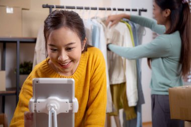 İki genç Asyalı kadın online giyim mağazasında yaşarken, evde sosyal medyada tablet ve dizüstü bilgisayarlı giysiler gösteriliyor. Girişimci çevrimiçi kıyafet satışı ve sunumu için çevrimiçi satış yapıyor..