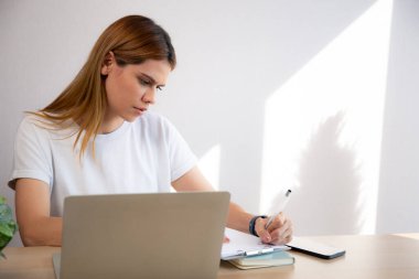 Ev ofisinin oturma odasındaki masanın üzerine not yazarken dizüstü bilgisayar kullanan genç iş kadını portresi, not defteri, girişimci ya da serbest meslek ve ticaret kullanan iş kadını.