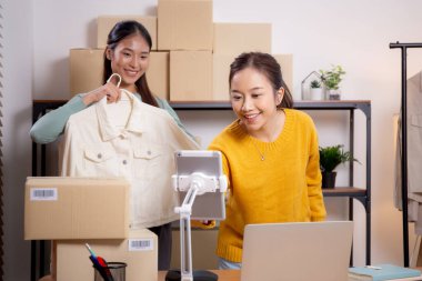 İki genç Asyalı kadın online giyim mağazasında yaşarken, evde sosyal medyada tablet ve dizüstü bilgisayarlı giysiler gösteriliyor. Girişimci çevrimiçi kıyafet satışı ve sunumu için çevrimiçi satış yapıyor..