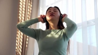 Portre takan, kulaklık takan, müzik dinleyen mutlu genç bir kadın. Evdeki oturma odasında eğleniyor ve eğleniyor. Mutluluk dolu bir kadın, tek bir kişi, yaşam tarzı kavramı..