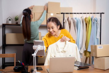 İki genç Asyalı kadın online giyim mağazasında yaşarken, evde sosyal medyada tablet ve dizüstü bilgisayarlı giysiler gösteriliyor. Girişimci çevrimiçi kıyafet satışı ve sunumu için çevrimiçi satış yapıyor..