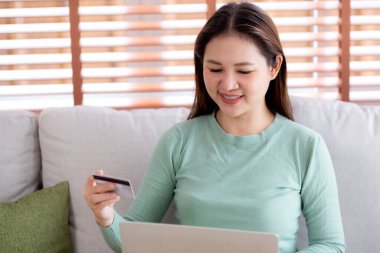 Kanepede oturan Asyalı genç bir kadın internet üzerinden dizüstü bilgisayar alışverişi yapıyor. İnternete kredi kartı alışverişi yapıyor. Kanepe, satın alma ve ödeme, iş ve yaşam tarzları ile kadın ödemesi yapıyor..