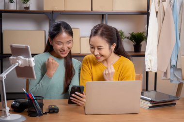 Başarı ve neşeli, girişimci veya çevrimiçi satıcı, ecommerce veya startup business, küçük işletme ile mağaza incelemesi için tablet ve dizüstü bilgisayar kullanan iki genç Asyalı kadın.