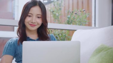Genç Asyalı iş kadını laptopuyla evde koltukta oturuyor, iş kadını evdeki not defterini kullanıyor, kadın dizüstü bilgisayarını kullanırken rahatlıyor başarı, iş konsepti.
