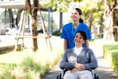 Genç Kafkas hemşire hastaya bakarken aynı zamanda tekerlekli sandalyeyi iterek parkta dinlenme ve rehabilitasyon, bakıcı desteği ve yardım hastası, sağlık ve sigorta, sağlık ve terapi.