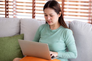 Genç Asyalı iş kadını portresi evdeki oturma odasında dizüstü bilgisayar kullanarak koltukta oturuyor, iş kadını kanepede oturuyor, rahat, iş ve serbest olarak defter üzerinde çalışıyor..