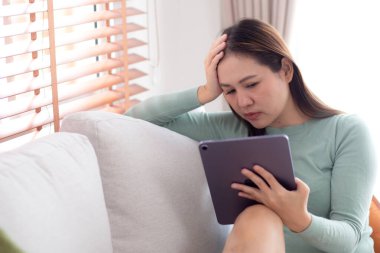 Tablet bilgisayar kullanan genç Asyalı iş kadını ev ofisinin oturma odasındaki kanepede otururken endişeli, iş kadını endişeli, yorgun, iş ve yaşam tarzını düşünürken tablet üzerinde çalışıyor..