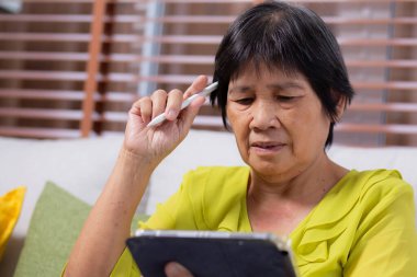 Kıdemli Asyalı kadın kanepeye oturmuş dijital tablet kullanıyor. Oturma odasında dinleniyor, yaşlılar tablete oturmuş düşünceli, rahat bir hayat tarzı ve iletişim kuruyor..