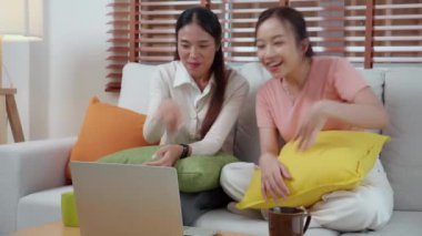 Kanepede oturan iki genç Asyalı kadın evdeki oturma odasında dizüstü bilgisayar kullanıyor, sosyal medyayı internet ve sohbet için kullanıyor, yaşam tarzı kavramı..
