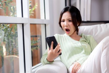 Asyalı genç bir kadın evdeki akıllı telefonuyla konuşurken koltukta oturmuş endişeli ve stresli bir şekilde oturuyor. Mutsuz ve endişeli bir kadın akıllı telefondan, yaşam tarzından ve iletişimden bahsediyor..