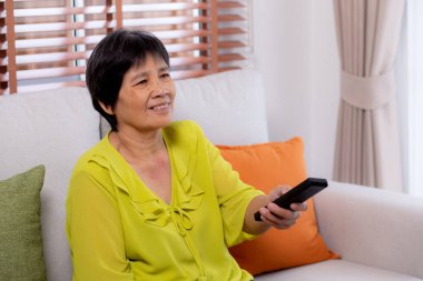 Kıdemli Asyalı kadın elinde oturma odasında televizyon seyretmek için uzaktan kumandayla koltukta oturuyor. Yaşlı kadın kanepede oturmuş, rahatlamış, yaşam tarzları ve eğlence tarzıyla televizyon seyrediyor..