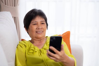 Üst düzey Asyalı kadın kanepeye oturmuş videolu telefon bekliyor. Oturma odasında. Yaşlılar sosyal medyayı internet üzerinden sohbet etmek için kullanıyor..