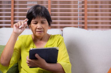 Kıdemli Asyalı kadın kanepeye oturmuş dijital tablet kullanıyor. Oturma odasında dinleniyor, yaşlılar tablete oturmuş düşünceli, rahat bir hayat tarzı ve iletişim kuruyor..