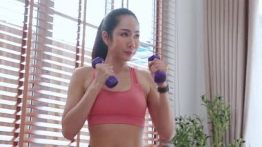 Spor giyinen genç Asyalı bir kadın evde güçlü ve güçlü olmak için halterlerle antrenman yapıyor kadın, kararlı bir şekilde antrenman yapıyor ve dambılları kaldırırken yumrukla mücadele ediyor..