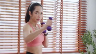 Spor giyinen genç Asyalı bir kadın evde güçlü ve güçlü olmak için halterlerle antrenman yapıyor kadın, kararlı bir şekilde antrenman yapıyor ve dambılları kaldırırken yumrukla mücadele ediyor..