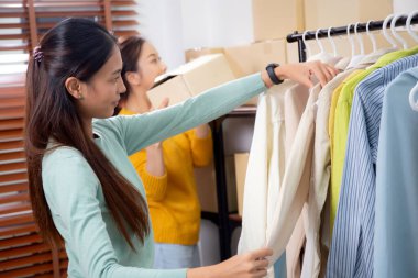 Hazırlık teslimatı için mağazada kıyafet düzenleyen iki genç Asyalı kadın, satıcı seçici giysiler, perakende satış için seçici giysiler, teslimat için mal, küçük işletme.