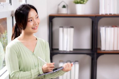 Genç Asyalı kadın, çalışma odasındaki oturma odasının penceresinin yanında ilham verici ve rahat bir şekilde tablet yazıyor. Çalışma alanı, iş yeri ve yaşam tarzında dijital tablet üzerinde çalışan neşeli bir kadın..