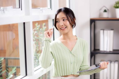 Genç Asyalı kadın tablet yazısını ilham kaynağı olarak kullanıyor ve ev ofisinin oturma odasının penceresinin yanında düşünceli bir şekilde dururken çalışma odasında dijital tablet üzerinde çalışan neşeli bir kadın, iş konsepti..