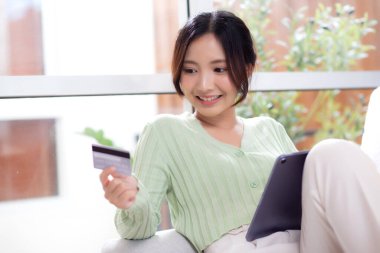 Kanepede oturan Asyalı genç bir kadın internet üzerinden tablet bilgisayar alışverişi yapıyor. İnternete kredi kartı alışverişi yapıyor. Neşeli bir kadın ise iş, satın alma ve ödeme, iş ve finans yapıyor..