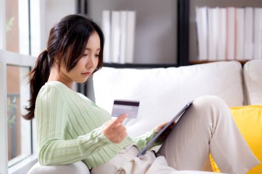 Genç Asyalı kadın, internet üzerinden tablet bilgisayar alışverişi yaparak kanepede oturuyor. Düşünceli, düşünceli, iş, ödeme, iş ve finans ile kadın ödemesi yapıyor..