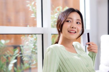 Kanepede oturan genç Asyalı kadın kredi kartı ya da banka kartı satın alırken düşünceli, düşünceli bir kadın iş yaparken, satın alma ve ödeme, iş ve finans.