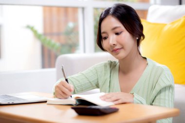 Evdeki oturma odasında ders kitabına yazarken finans ve yatırım veya vergi hakkında planlama ve strateji yapan genç Asyalı kadın, ev, iş ve finans giderleri ve bütçesi olan kadın.