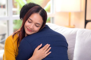 Genç Asyalı çift, evdeki oturma odasında sevgi dolu bir şekilde kanepede otururken birbirlerine sarılırlar. Karı koca, nazik ve sevgi dolu bir şekilde birbirlerine sarılırlar. Birbirlerine sarılırlar..