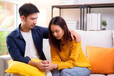 Asyalı genç adam, üzgün kadını nazikçe kucaklayarak rahatlatıyor. Oturma odasındaki kanepede oturan kadın, ifadesiyle mutsuz ve endişeli bir eş. Kocası ise şefkat ve yakınlıkla teselli ediyor..