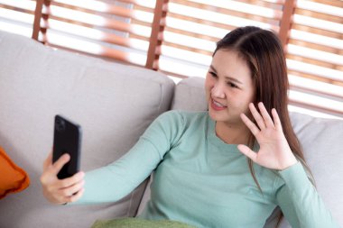 Genç Asyalı bir kadın kanepede oturmuş videolu telefon bekliyor. Oturma odasında telefon görüşmesi yapıyor. Sosyal medyayı internet üzerinden sohbet etmek için kullanıyor..