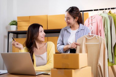 İki genç Asyalı iş kadını dizüstü bilgisayar, sevkiyat ve kargo için müşteri kıyafetlerini kontrol ediyor, çevrimiçi satıcı ve mağaza ile perakende satış, başlangıç veya küçük işletme, nakliye ve envanter.