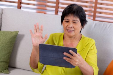 Üst düzey Asyalı kadın dijital tablette el sallıyor oturma odasındaki koltukta oturuyor, yaşlılara ait video görüntüsü tablette, koltukta oturuyor, emeklilik ve teknoloji..
