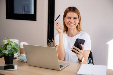 Kredi kartı ya da banka kartı tutan mutlu beyaz kadın akıllı telefon kullanırken internet üzerinden alışveriş yaparak ödeme yapıyor, finans, iş ve mali işlemler yaparak harcama yapan kadın.