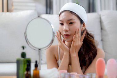 Güzel Asyalı bir kadın, oturma odasında aynaya bakarken kırışıklıklar için yüzüne krem sürüyor. Güzel bir kadın, yanağına nemlendirici sürüyor. Hijyen, cilt bakımı ve güzellik için..