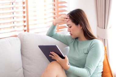 Tablet bilgisayar kullanan genç Asyalı iş kadını ev ofisinin oturma odasındaki kanepede otururken endişeli, iş kadını endişeli, yorgun, iş ve yaşam tarzını düşünürken tablet üzerinde çalışıyor..