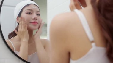 Genç ve güzel Asyalı bir kadın banyoda aynaya bakarken cildine krem sürüyor ya da losyon sürüyor. Tedavi için nemlendirici sürüyor. Yüz hatları, güzellik ve cilt bakımı için..