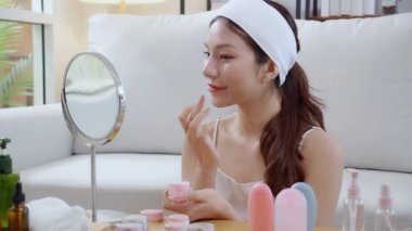 Güzel Asyalı bir kadın, oturma odasında aynaya bakarken kırışıklıklar için yüzüne krem sürüyor. Güzel bir kadın, yanağına nemlendirici sürüyor. Hijyen, cilt bakımı ve güzellik için..