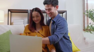 Genç Asyalı çift rahat ve rahat bir şekilde dizüstü bilgisayara bakıyor. Oturma odasındaki kanepede oturuyor. Kanepede dizüstü bilgisayar izliyor..