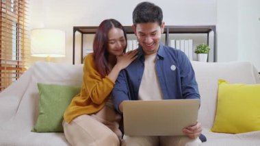 Genç Asyalı çift rahat ve rahat bir şekilde dizüstü bilgisayara bakıyor. Oturma odasındaki kanepede oturuyor. Kanepede dizüstü bilgisayar izliyor..