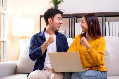 Genç Asyalı çift rahat ve başarı için dizüstü bilgisayara bakıyor. Oturma odasındaki kanepede oturuyor. Kanepede dizüstü bilgisayar izliyor..