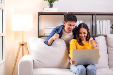 Genç Asyalı çift rahat ve başarı için dizüstü bilgisayara bakıyor. Oturma odasındaki kanepede oturuyor. Kanepede dizüstü bilgisayar izliyor..