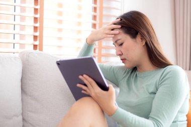 Tablet bilgisayar kullanan genç Asyalı iş kadını ev ofisinin oturma odasındaki kanepede otururken endişeli, iş kadını endişeli, yorgun, iş ve yaşam tarzını düşünürken tablet üzerinde çalışıyor..