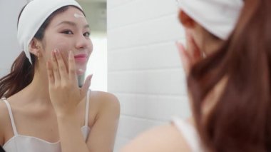 Genç ve güzel Asyalı bir kadın banyoda aynaya bakarken cildine krem sürüyor ya da losyon sürüyor. Tedavi için nemlendirici sürüyor. Yüz hatları, güzellik ve cilt bakımı için..
