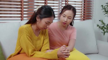 Genç Asyalı bir kadın, arkadaşını ağlama ve depresyonla rahatlatırken oturma odasında kanepede oturuyor, arkadaşlığın sıkıntısı ve empatisi, umutsuzluk ve ruh sağlığını desteklerken, bir kadın da arkadaşını teşvik ediyor..