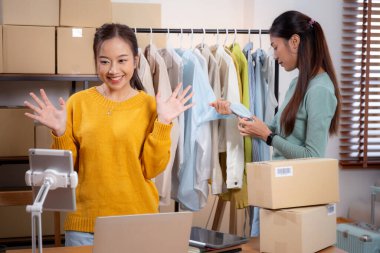 İki genç Asyalı kadın online giyim mağazasında yaşarken, evde sosyal medyada tablet ve dizüstü bilgisayarlı giysiler gösteriliyor. Girişimci çevrimiçi kıyafet satışı ve sunumu için çevrimiçi satış yapıyor..